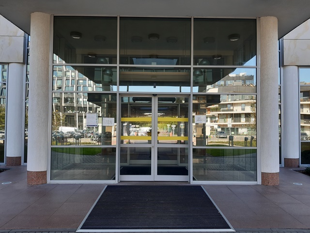 Zdjęcie przedstawia wejście główne do budynku. Szklane, automatycznie otwierane drzwi są oznaczone poziomymi żółtymi pasami ostrzegawczymi. Nad drzwiami znajduje się nadajnik systemu Totupoint dla osób słabowidzących i niewidomych.