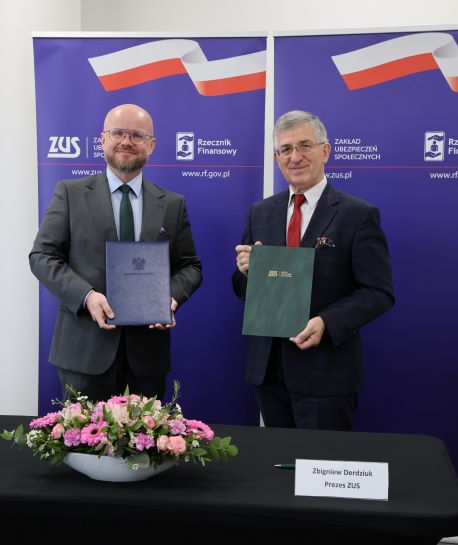Dwóch mężczyzn, rzecznik finansowy i prezes ZUS stoją przy stole. W ręku trzymają teczki z podpisanym porozumieniem i współpracy.