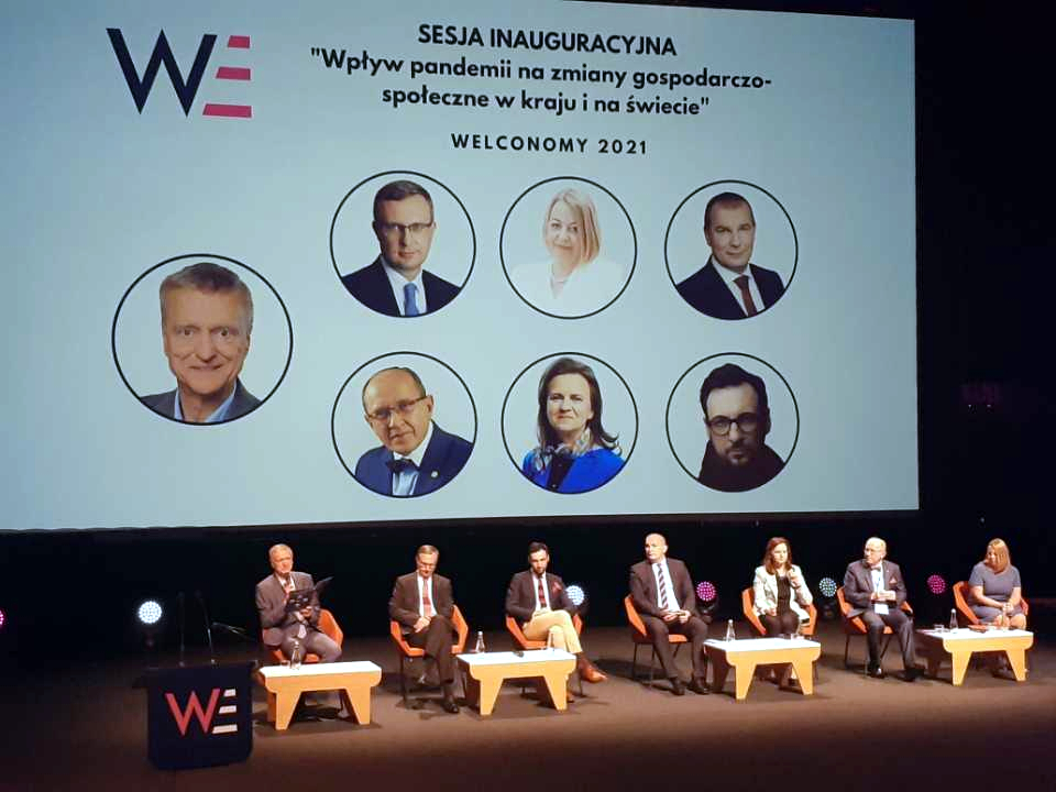 Welconomy Forum w Toruniu - sesja inauguracyjna. paneliści siedzą w rzędzie na tle ekranu. 6 panelistów i i porwadzący, na ekranie okrągłe zdjęcia panelistów i tytuł sesji
